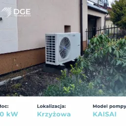 Zewnętrzna jednostka pompy ciepła Kaisai zamontowana przy ścianie budynku, widoczna zza krzewów.