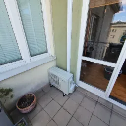 Kompaktowa jednostka klimatyzacji zewnętrznej, marki SEVRA, umieszczona na balkonie z terakotową podłogą, obok doniczki z uschniętą rośliną i otwartych drzwi balkonowych z widokiem na wnętrze...