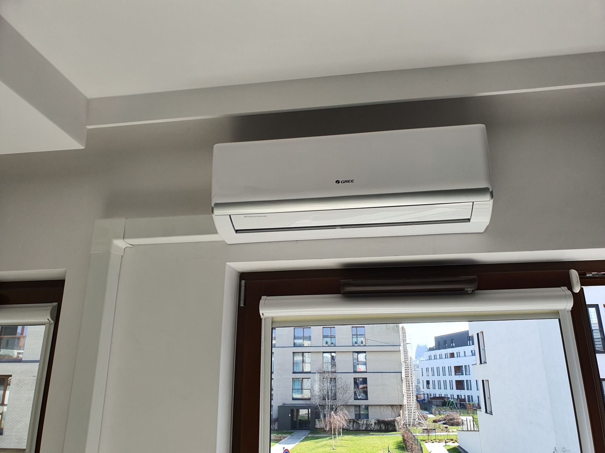 Biały klimatyzator marki Gree zamontowany na ścianie nad oknem z roletą, widok na osiedle z zielonym trawnikiem.
