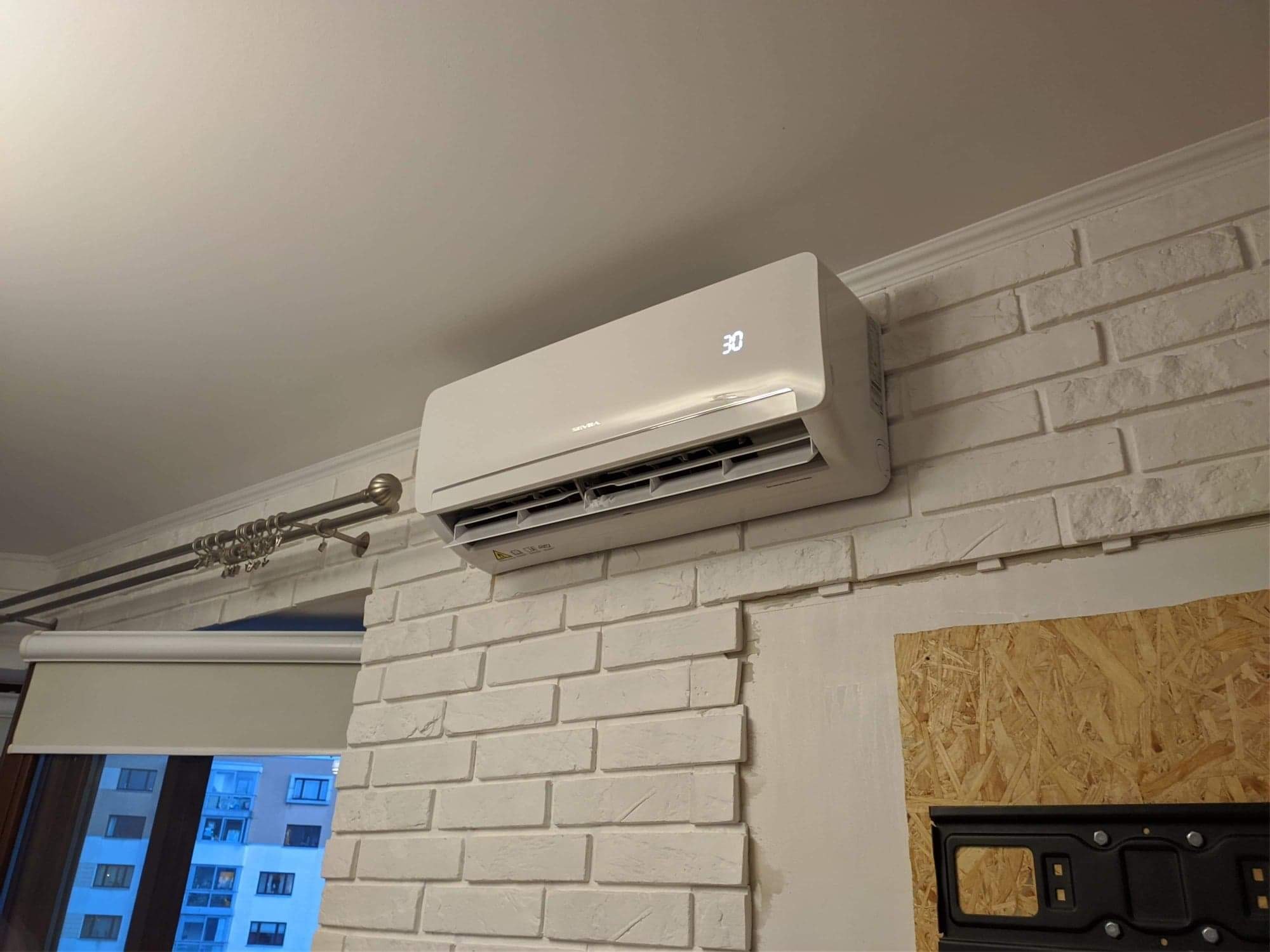 Biała jednostka klimatyzacji ściennej z wyświetlaną temperaturą 30 stopni, zamontowana na ścianie z białej cegły w mieszkaniu z widokiem na inne budynki za oknem.