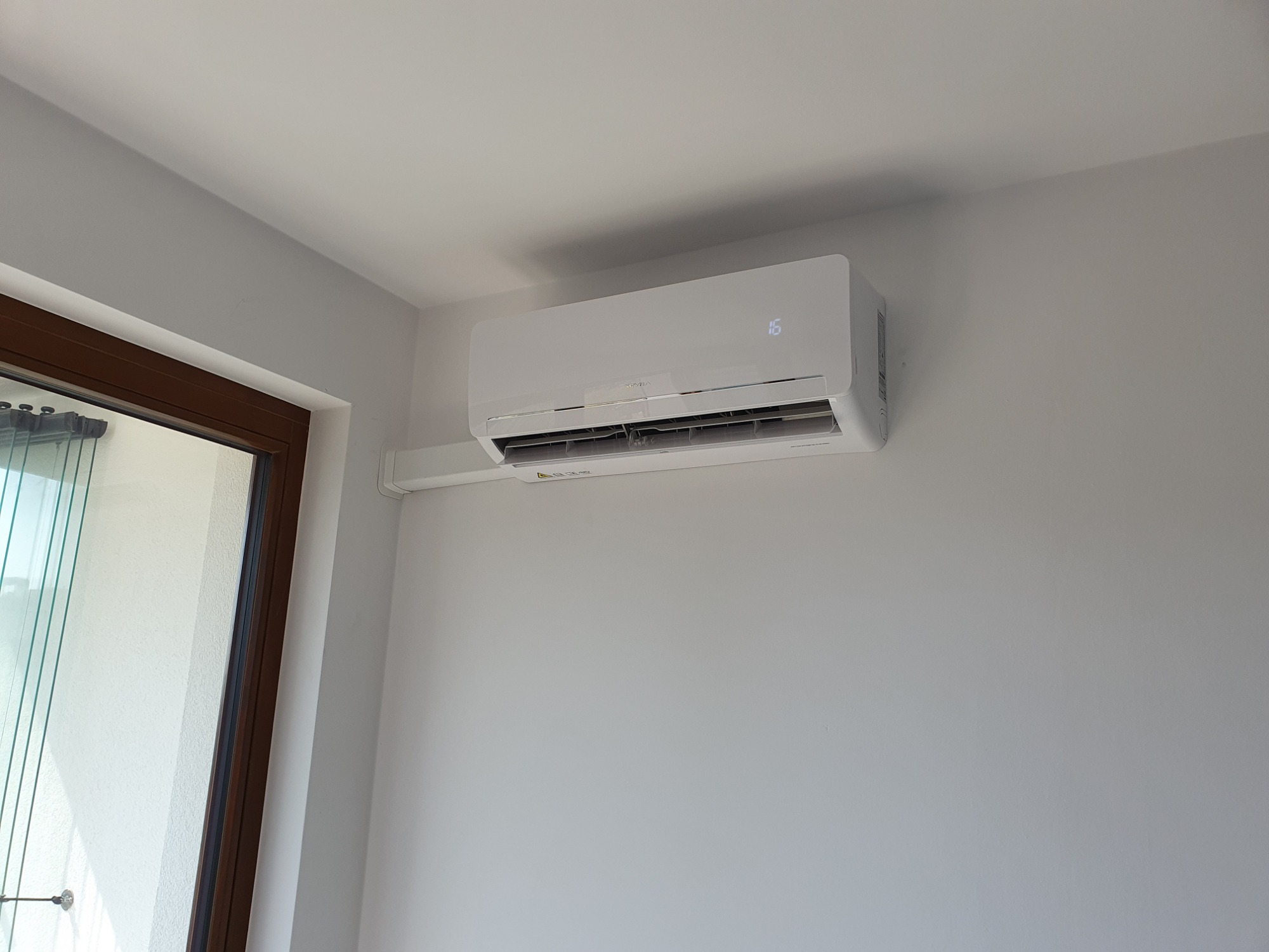 Biała klimatyzacja ścienna z wyświetlaczem temperatury 16 stopni, zamontowana w jasnym pomieszczeniu z oknem balkonowym.