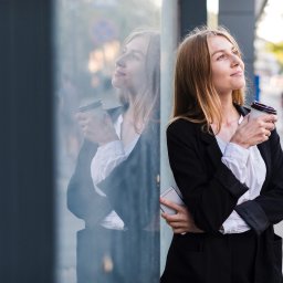 Akademia Kobiecej Przedsiębiorczości - Kobieta w marynarce, z kawą, oparta o szklaną ścianę budynku. Widoczne odbicie i smartfon w ręku. Uśmiechnięta, patrzy w górę. Zieleń drzew w tle.