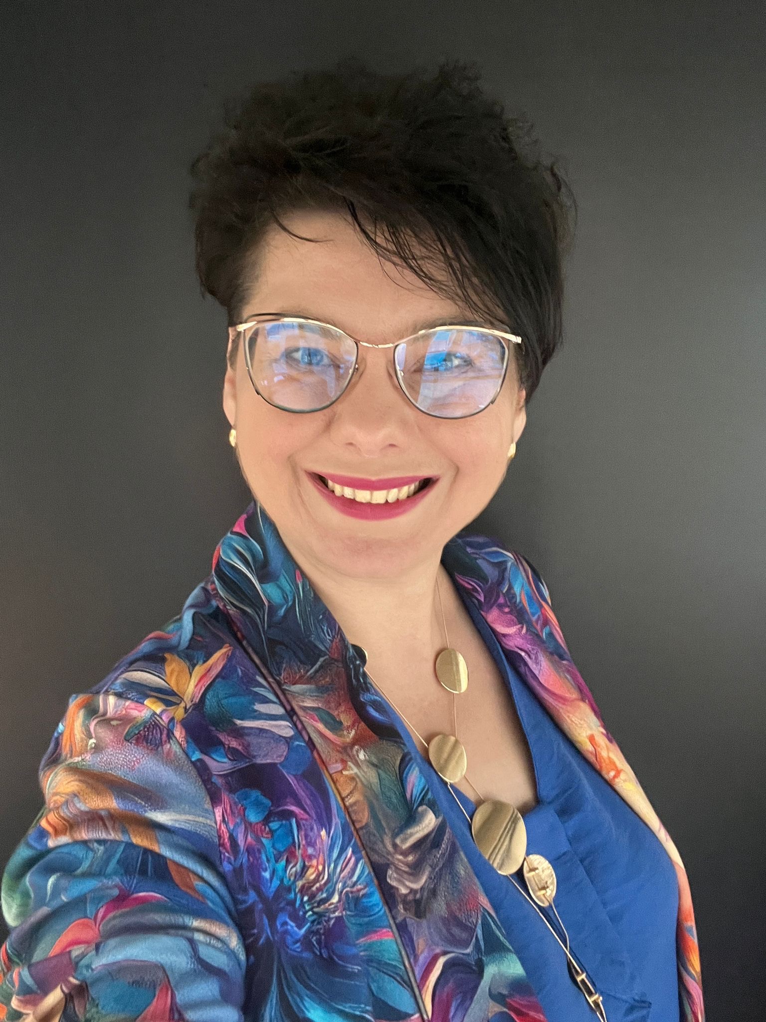 Uśmiechnięta kobieta w okularach i wzorzystej marynarce na ciemnoszarym tle. Widoczny złoty naszyjnik i niebieska bluzka.