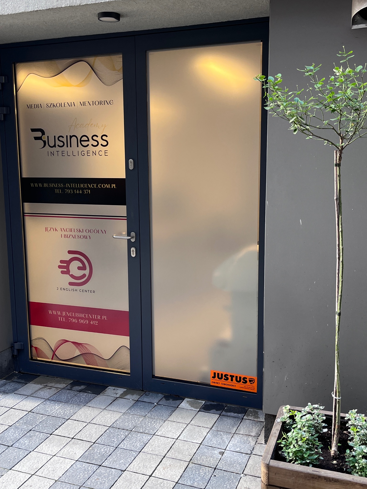 Wejście do biura z naklejkami informacyjnymi: Akademia Business Intelligence i J English Center. Obok donica z drzewkiem.