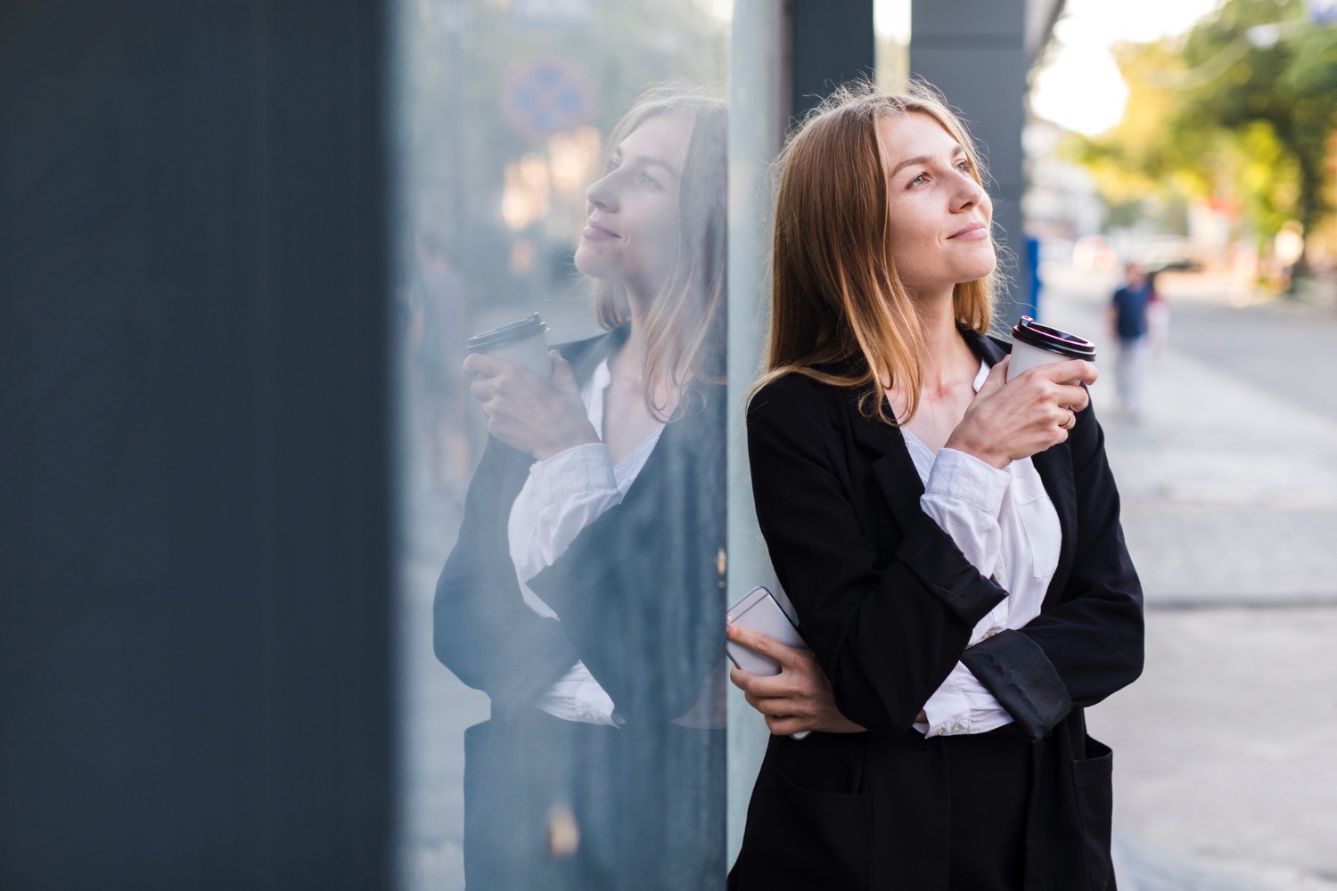 Kobieta w marynarce, z kawą, oparta o szklaną ścianę budynku. Widoczne odbicie i smartfon w ręku. Uśmiechnięta, patrzy w górę. Zieleń drzew w tle.