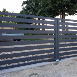 Nowoczesna, dwuskrzydłowa brama wjazdowa z poziomych, grafitowych profili, zamontowana na słupkach z szarego, łupanego kamienia. W tle widoczny zielony trawnik i drzewa.