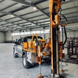 Mobilna wiertnica zamontowana na srebrnym pick-upie w hali produkcyjnej, podczas wiercenia otworu w posadzce betonowej. Widoczne hydrauliczne podpory stabilizujące maszynę.