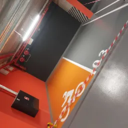 Wnętrze parkingu podziemnego z pomarańczową posadzką i szarą nawierzchnią z wymalowanym symbolem roweru, widoczne rury wentylacyjne pod sufitem i instalacja przeciwpożarowa.