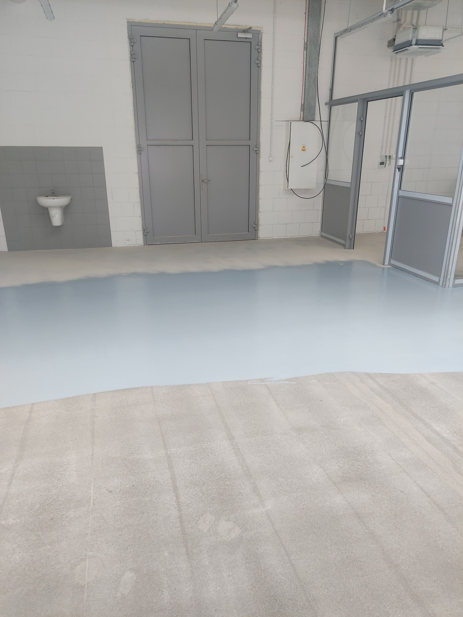 Wylewka samopoziomująca w jasnym odcieniu szarości, częściowo rozlana na betonowej podłodze w przemysłowym wnętrzu z szarymi drzwiami i umywalką w tle.