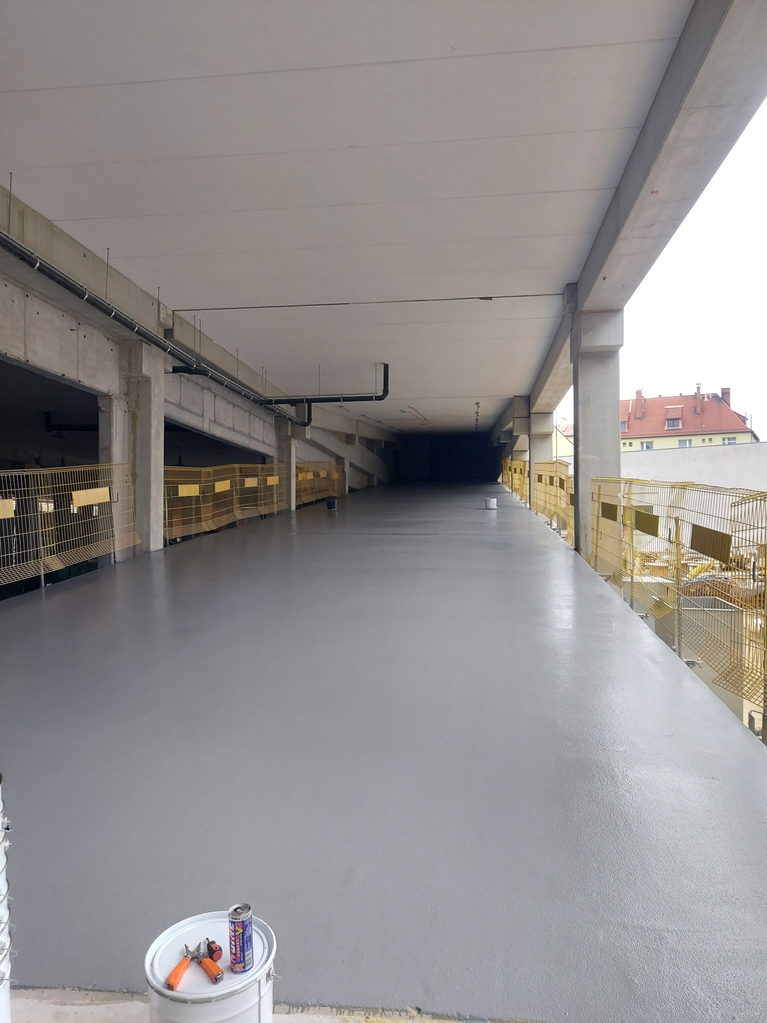 Świeżo wylana, szara powierzchnia na parkingu wielopoziomowym, zabezpieczona żółtymi barierkami, z widocznym wiadrem farby i puszką napoju energetycznego w rogu.