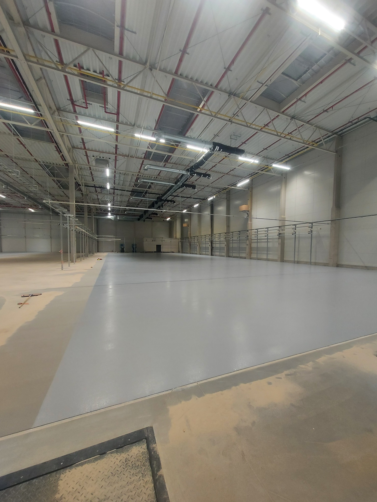 Szara, gładka posadzka żywiczna w hali magazynowej, widoczne instalacje pod sufitem i metalowe barierki wzdłuż ściany.