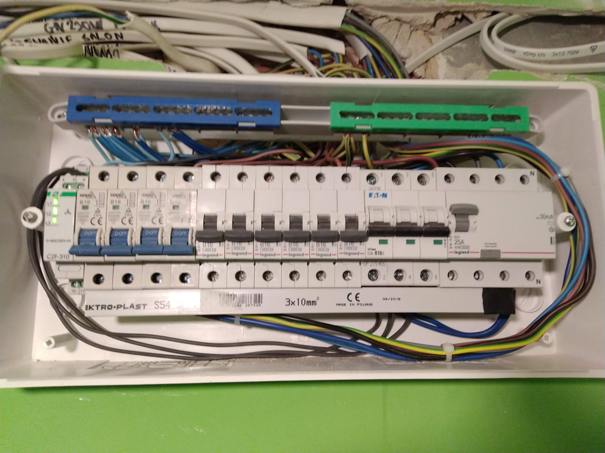 Otwarta skrzynka elektryczna z zainstalowanymi bezpiecznikami EATON i Legrand, widoczne kable w różnych kolorach, listwy zaciskowe niebieskie i zielone, oraz oznaczenia na przewodach. W tle...