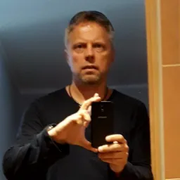 Mężczyzna robi selfie w lustrze łazienkowym, trzymając czarny smartfon, widoczne odbicie w lustrze i fragment pomarańczowej ramy drzwi.