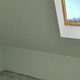 Poddasze po malowaniu na szaro, widoczne okno dachowe z drewnianą ramą i surowa betonowa podłoga.