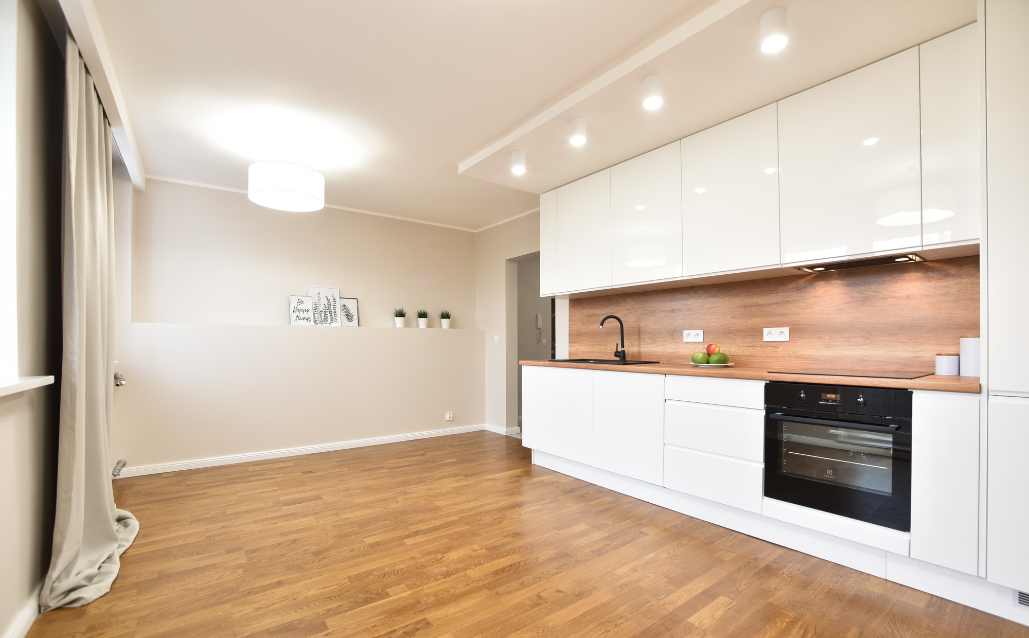 Jasne wnętrze z drewnianą podłogą, białą zabudową kuchenną z drewnianym blatem i czarną armaturą, minimalistyczne dekoracje na ścianie, całość oświetlona ciepłym światłem.