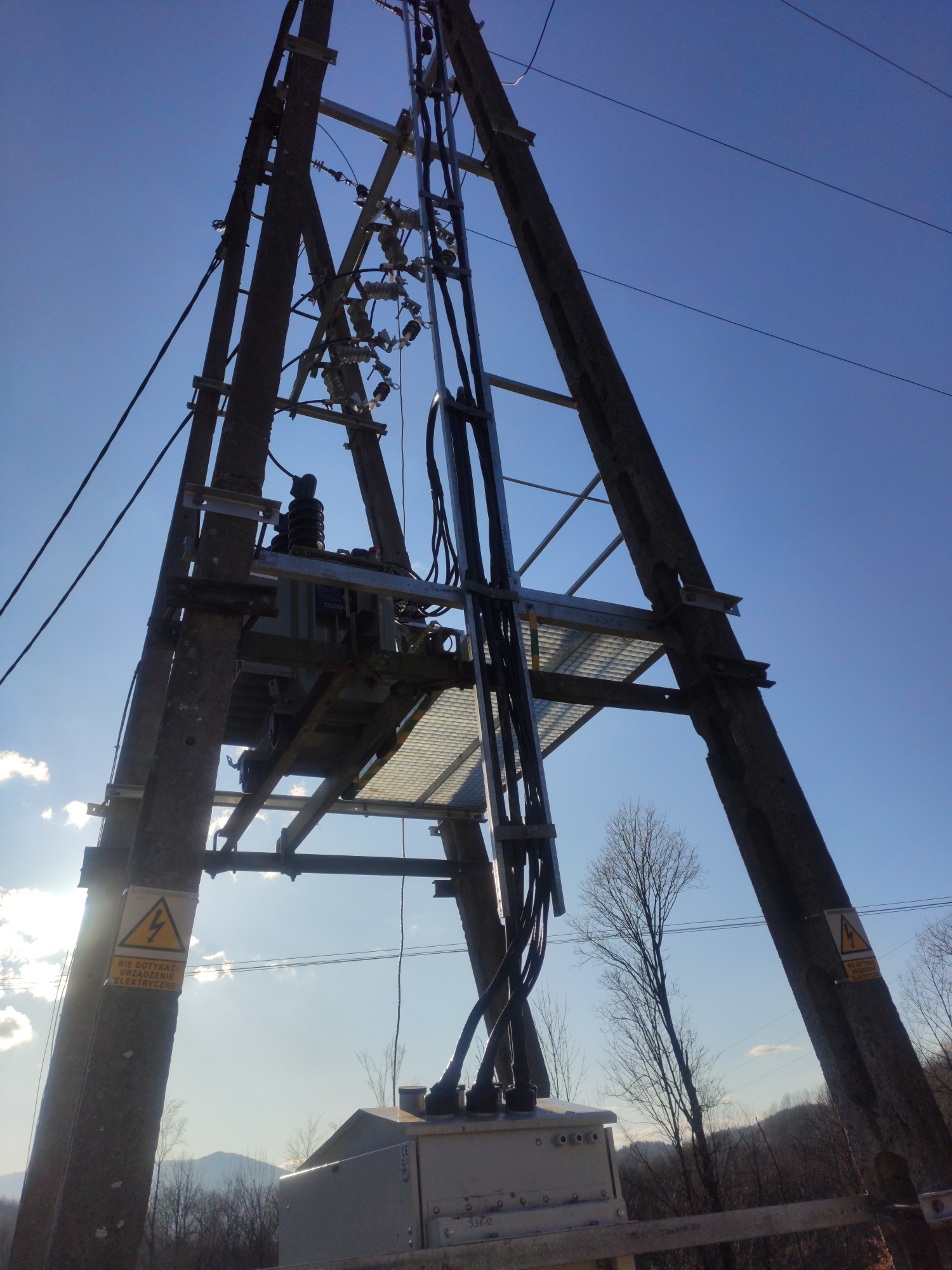 Słup energetyczny z transformatorem i przewodami, widoczne znaki ostrzegawcze, na tle błękitnego nieba i zalesionych wzgórz.