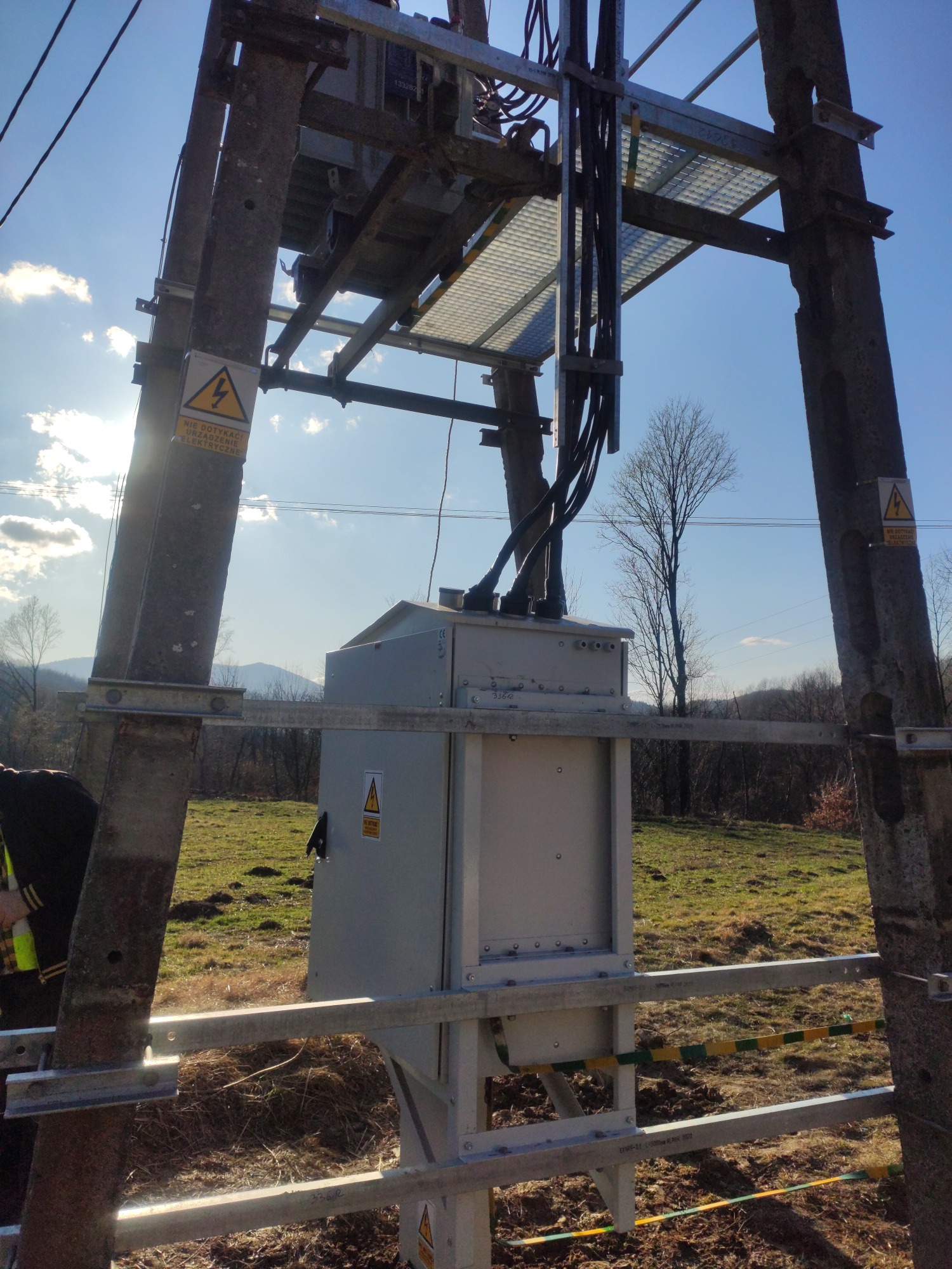 Słup elektryczny z zamontowaną skrzynką rozdzielczą i platformą, widoczne kable, tabliczki ostrzegawcze i krajobraz w tle.
