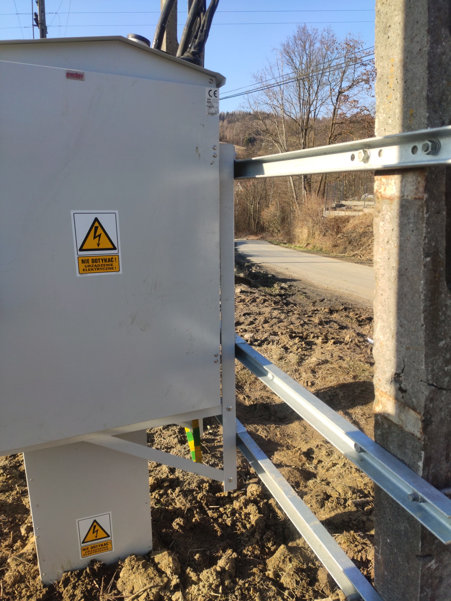 Szara skrzynka elektryczna z ostrzeżeniem 'Nie dotykać! Urządzenie elektryczne!' zamontowana na metalowych profilach przy betonowym słupie, widok na drogę i wzgórza w tle.