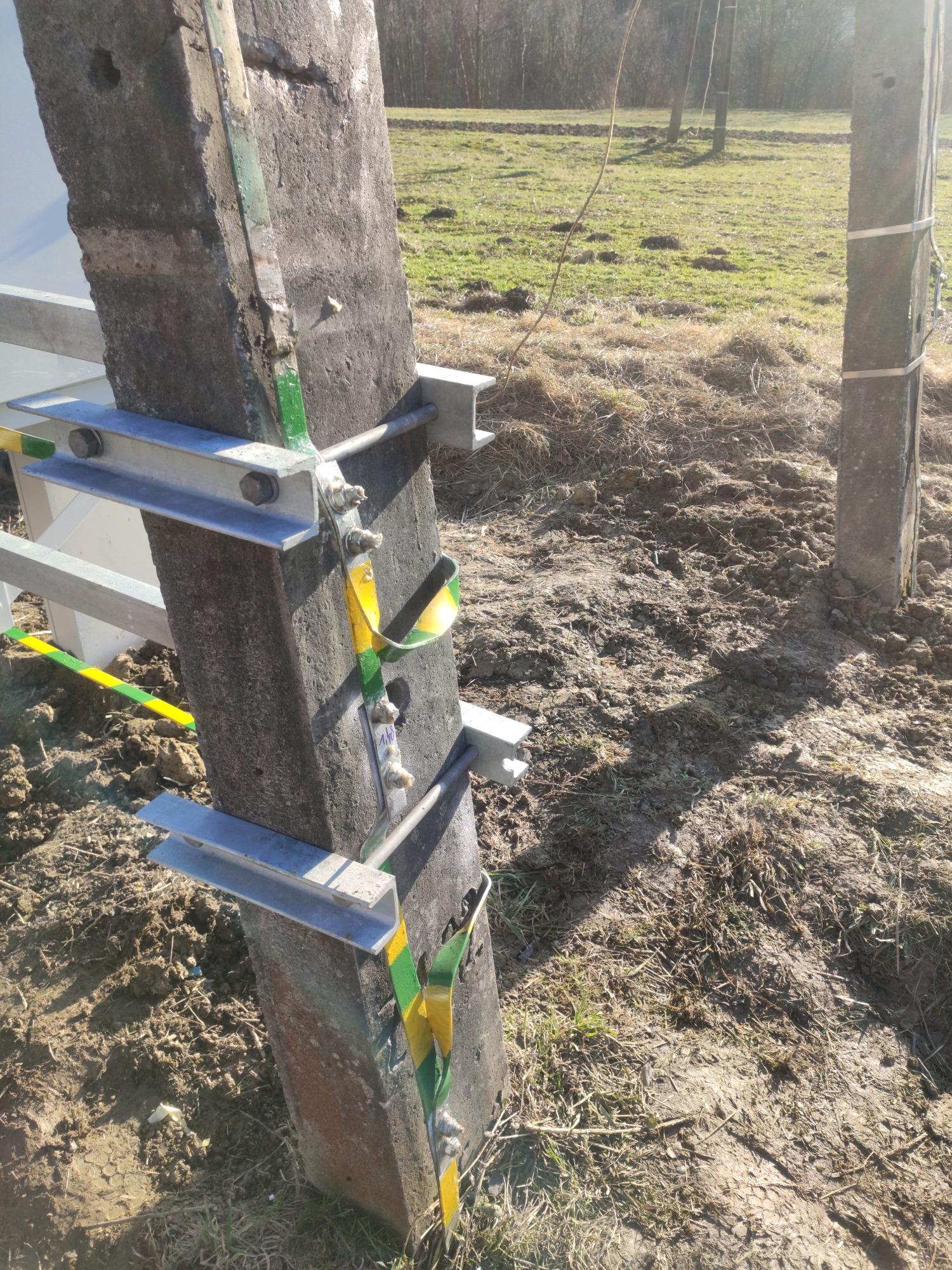 Betonowy słup z zamontowanymi metalowymi wspornikami i taśmą ostrzegawczą żółto-zieloną, na tle ziemi z resztkami trawy. Widoczne kolejne słupy w oddali.