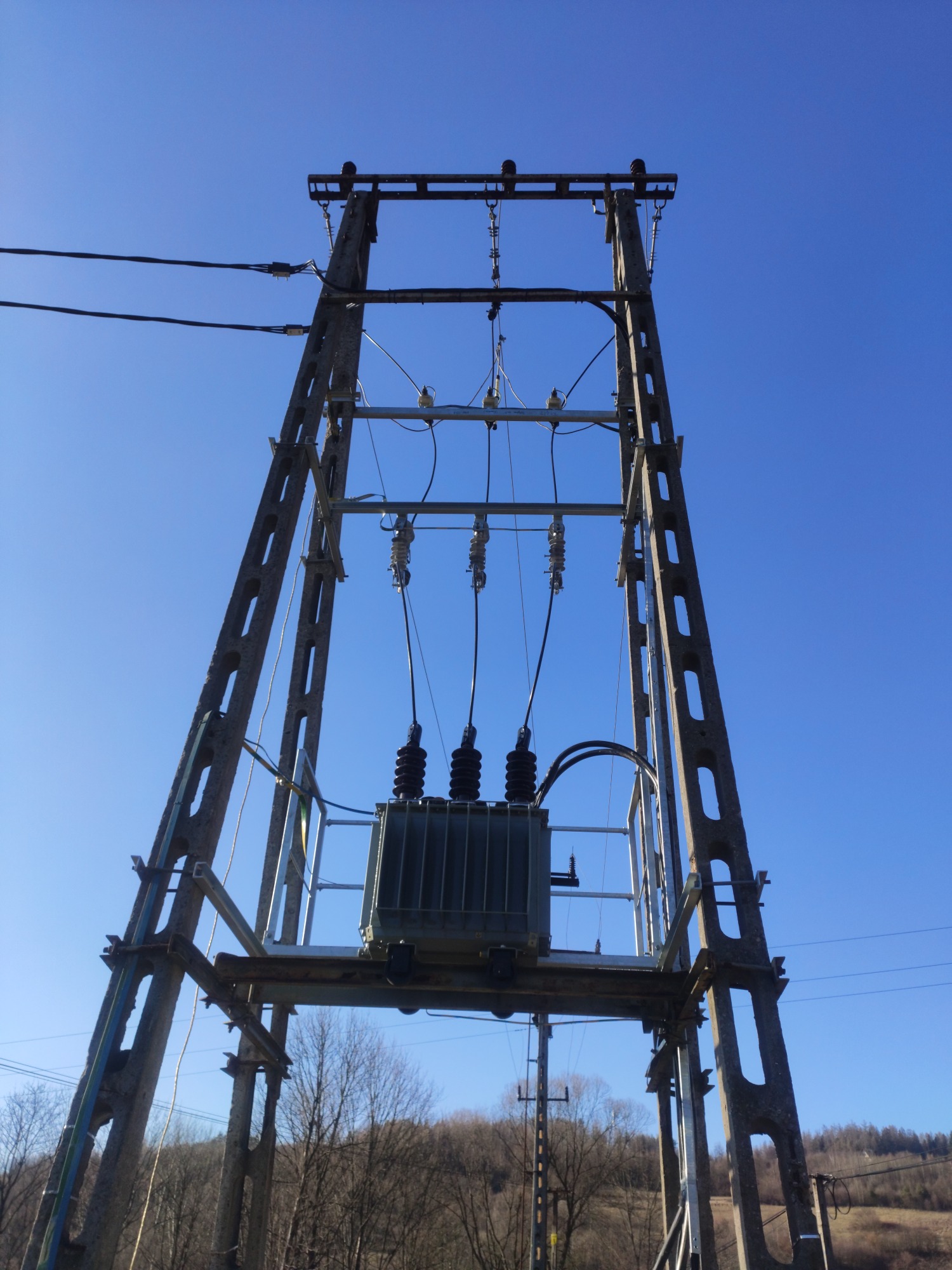 Stacja transformatorowa na betonowym słupie kratownicowym, widok z dołu na tle błękitnego nieba i drzew bez liści.