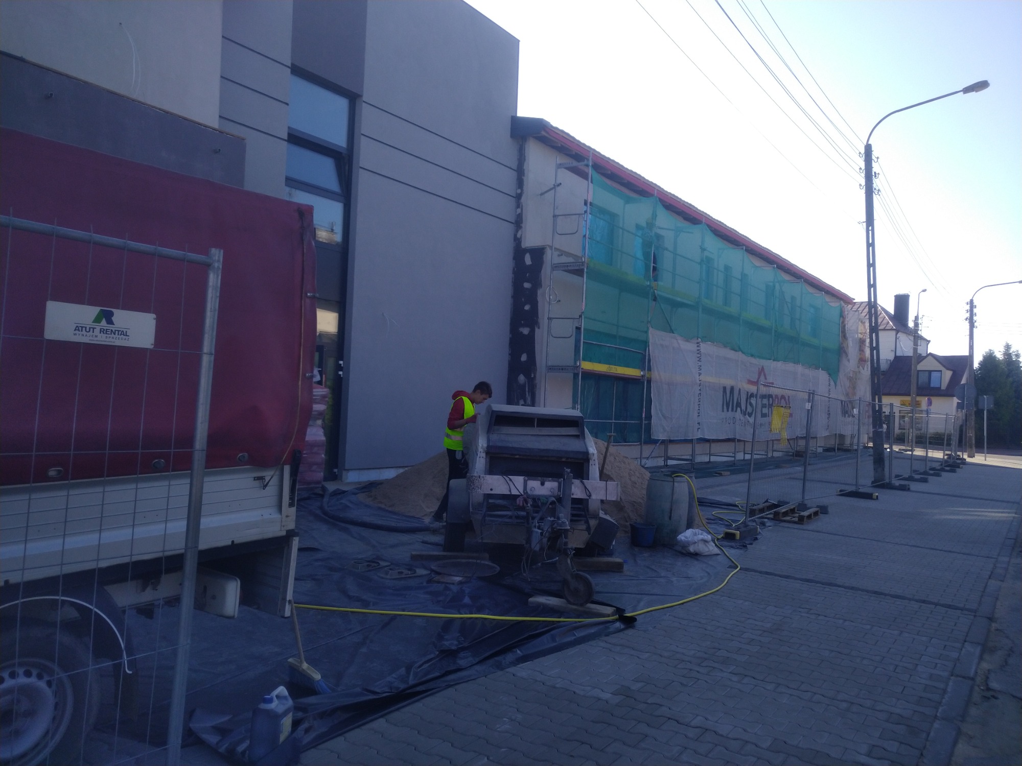 Pracownik w kamizelce odblaskowej obsługuje maszynę do przygotowywania mieszanki na placu budowy, obok zaparkowany samochód dostawczy z widocznym logo firmy wynajmującej sprzęt budowlany, w tle...