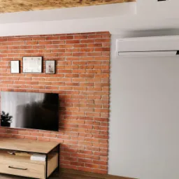 Biała klimatyzacja Samsung zamontowana na szarej ścianie w pokoju z ceglaną ścianą i drewnianym sufitem.