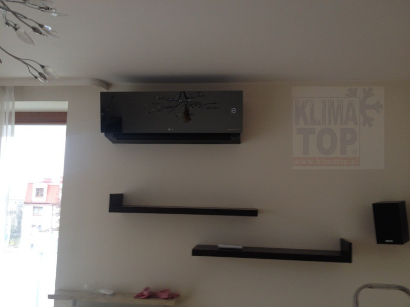 Czarny klimatyzator LG na ścianie w pokoju z półkami i logiem firmy instalującej, widoczne ustawienie temperatury na wyświetlaczu.