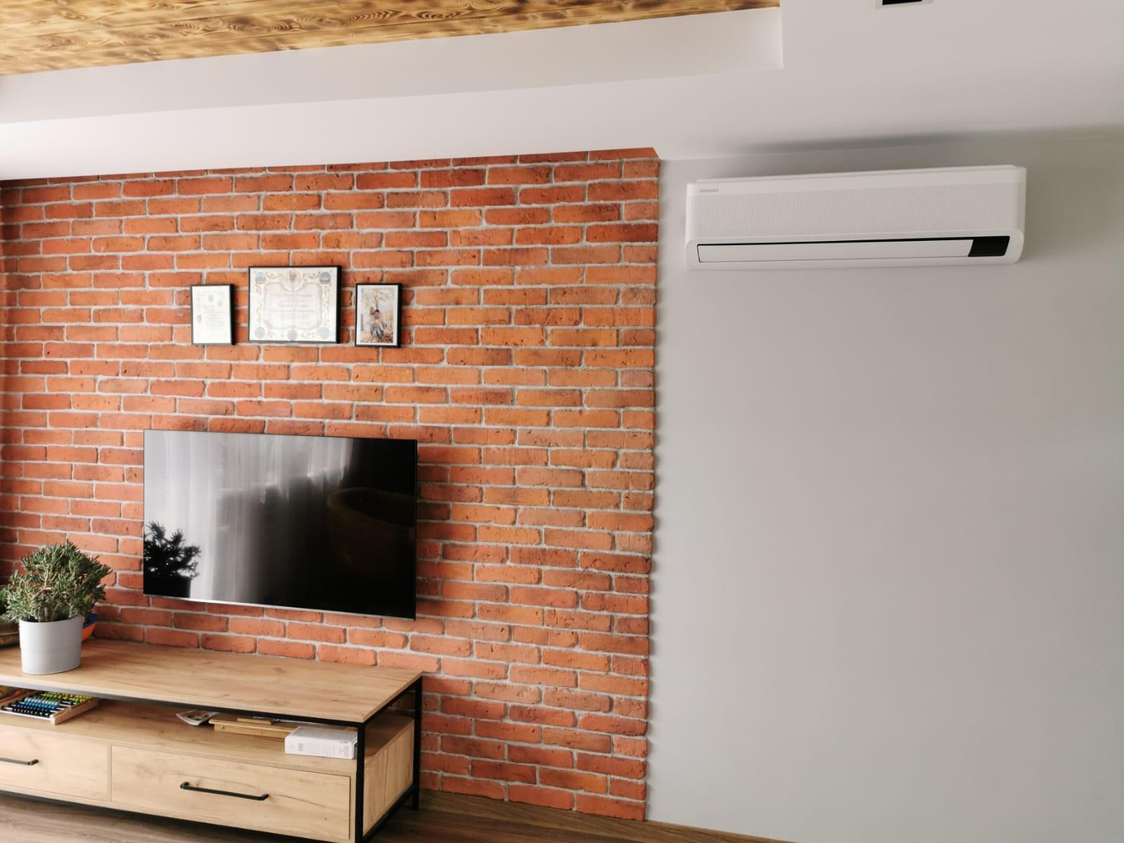 Biała klimatyzacja Samsung zamontowana na szarej ścianie w pokoju z ceglaną ścianą i drewnianym sufitem.