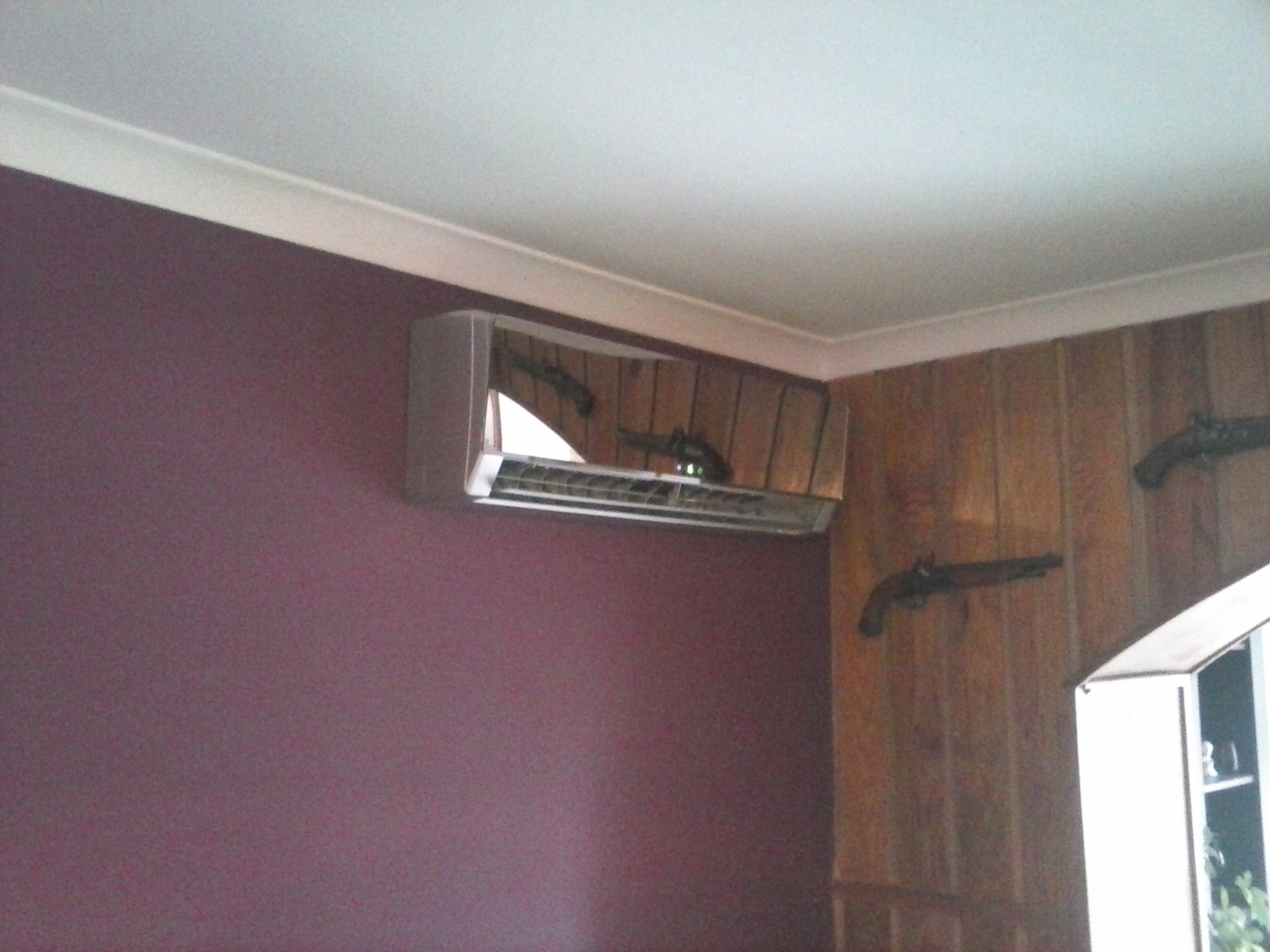 Zamontowana klimatyzacja ścienna w pomieszczeniu z bordową ścianą i boazerią, na której wiszą dekoracyjne repliki pistoletów.