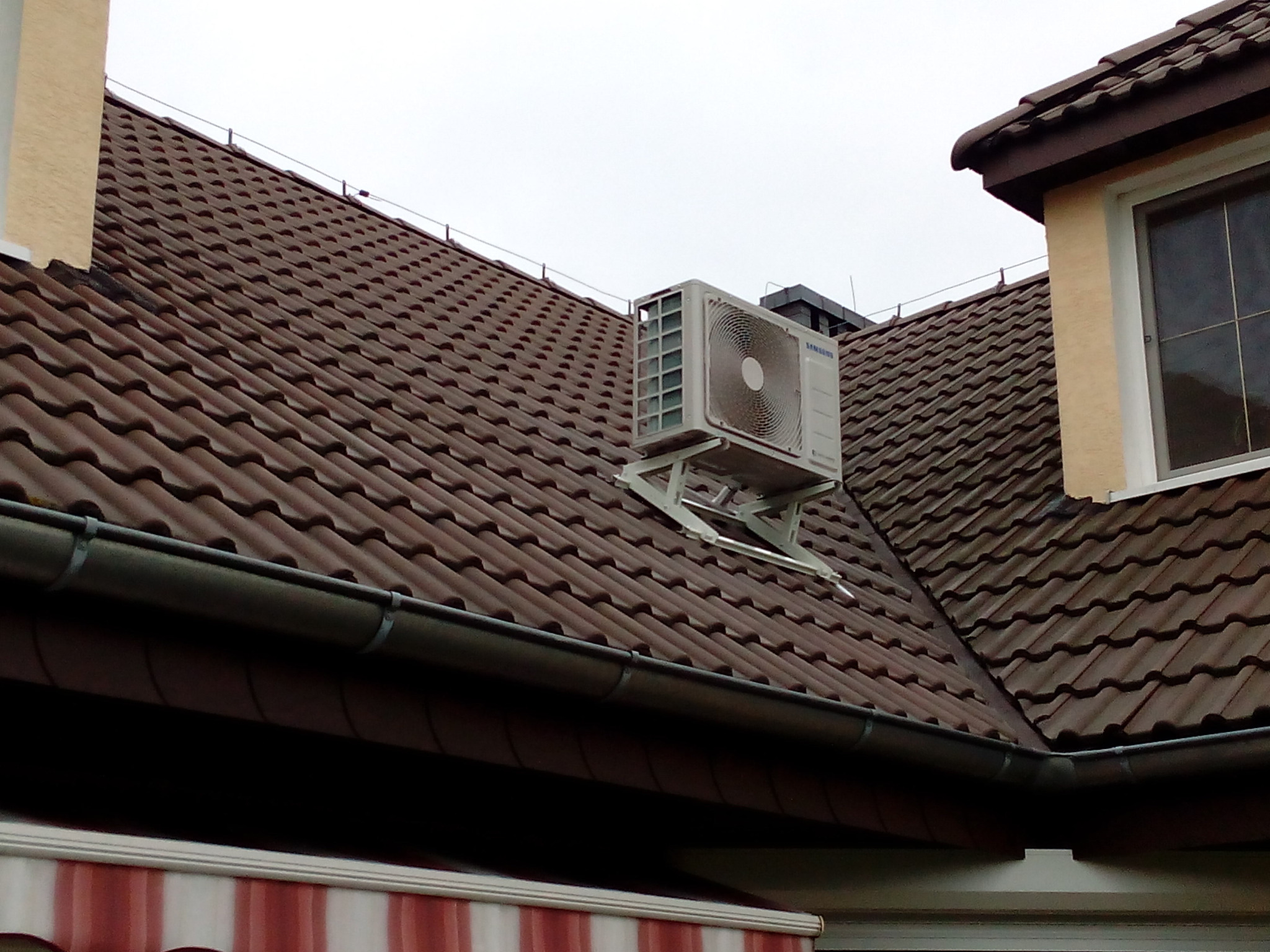 Biała jednostka zewnętrzna klimatyzacji Samsung zamontowana na specjalnej metalowej konstrukcji na brązowym dachu domu z dachówki ceramicznej.