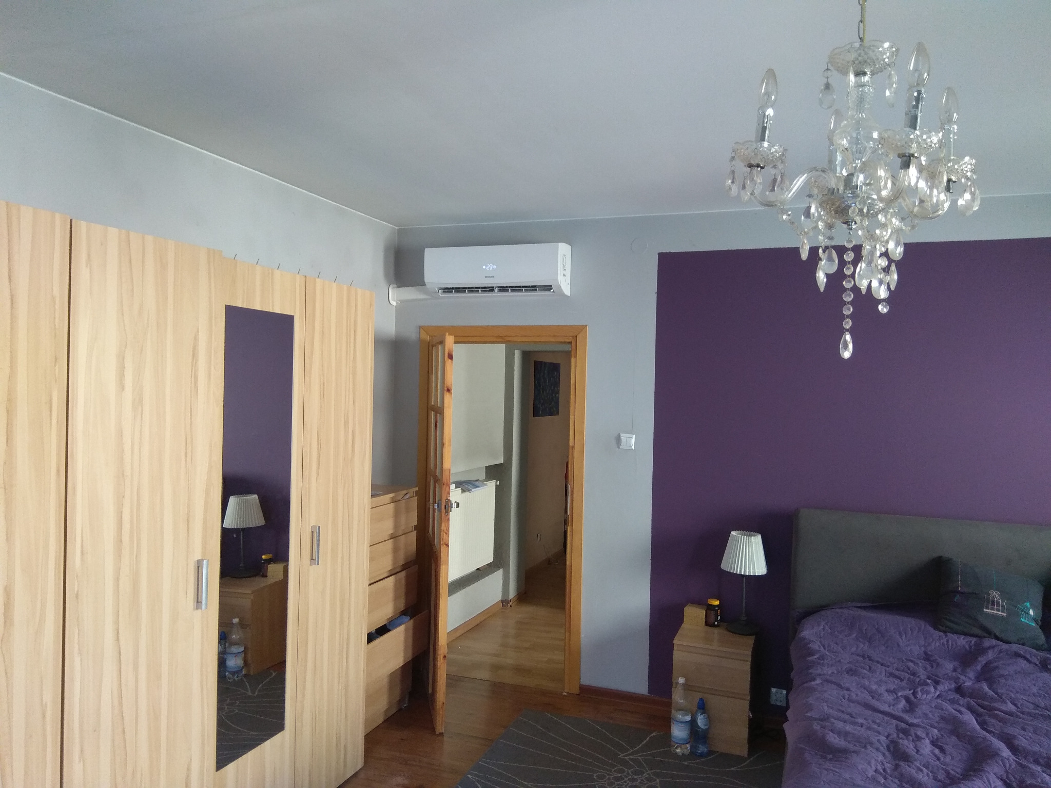 Klimatyzator ścienny zamontowany w sypialni z fioletową ścianą, widoczny nad drzwiami, obok szafy i żyrandola.