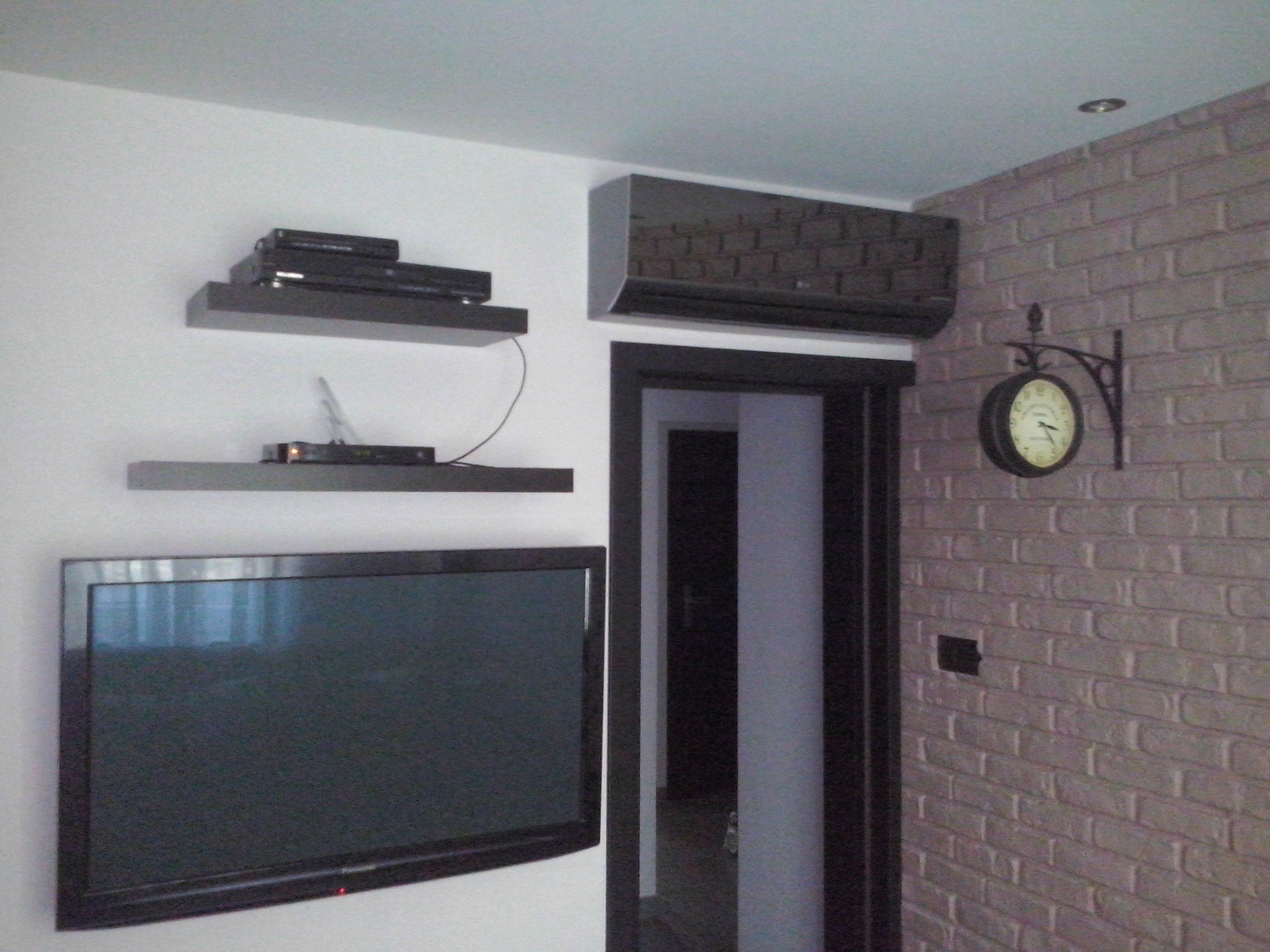Czarny klimatyzator ścienny w nowoczesnym salonie z telewizorem, półkami i zegarem na ścianie z cegły dekoracyjnej.
