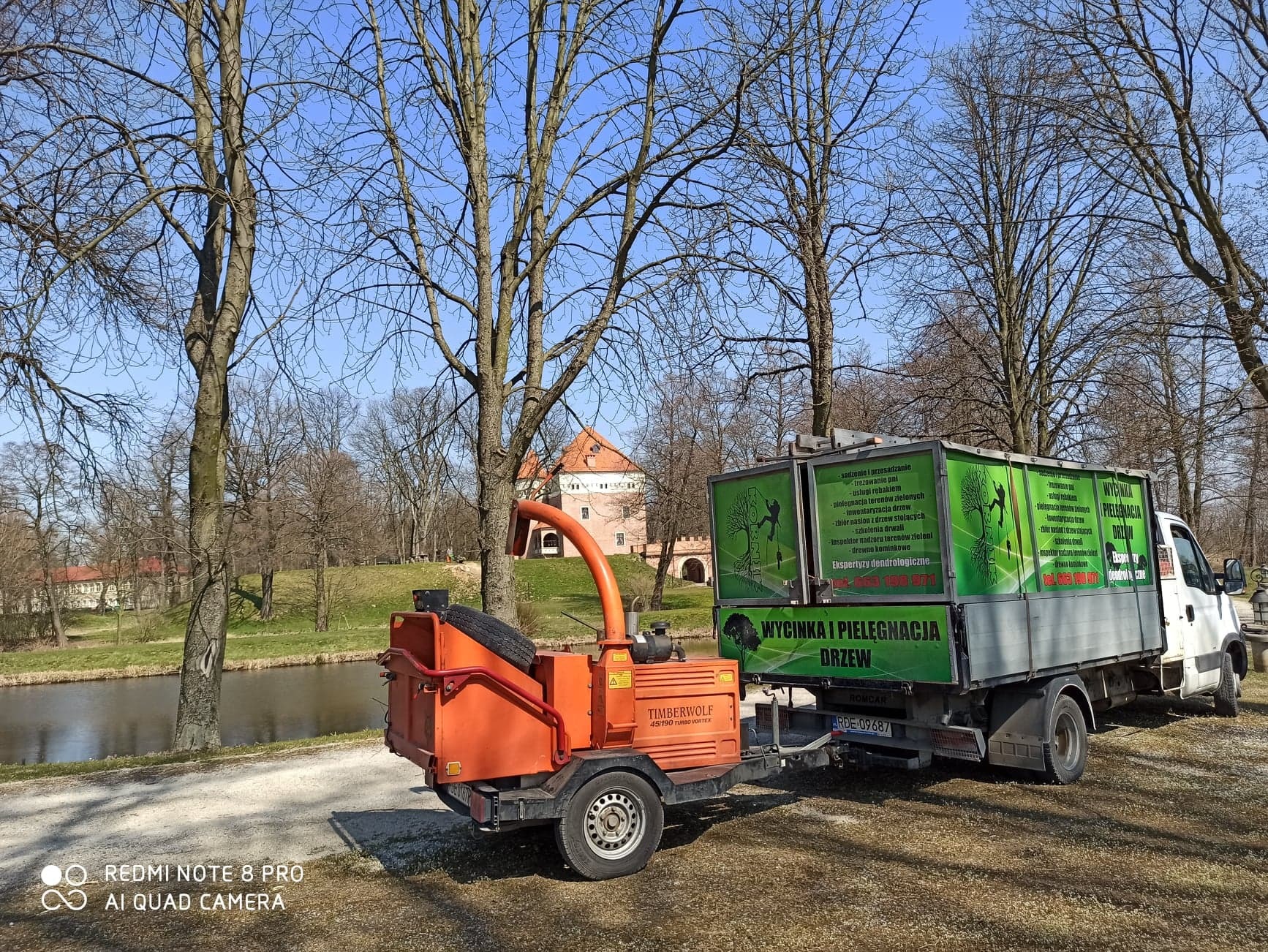 Pomarańczowy rębak do gałęzi Timberwolf podpięty do białej ciężarówki z reklamą usług związanych z drzewami, na tle parku z zamkiem i stawem w słoneczny dzień.