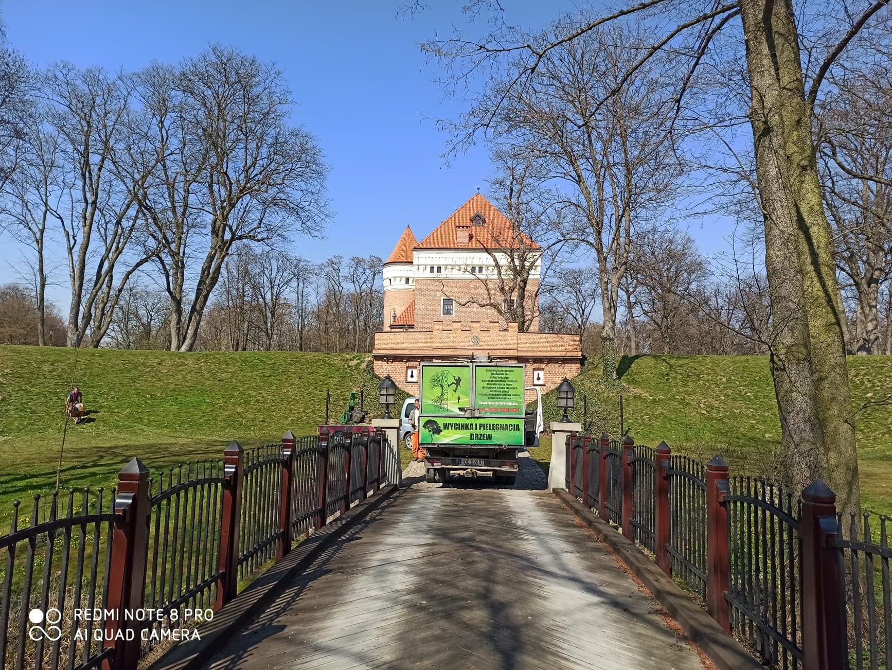Widok z mostu na zamek, gdzie w tle stoi zielony samochód z napisem 'Wycinka i pielęgnacja drzew' oraz pracownik w pomarańczowym ubraniu, a z lewej strony w oddali widać osobę pracującą z długim...