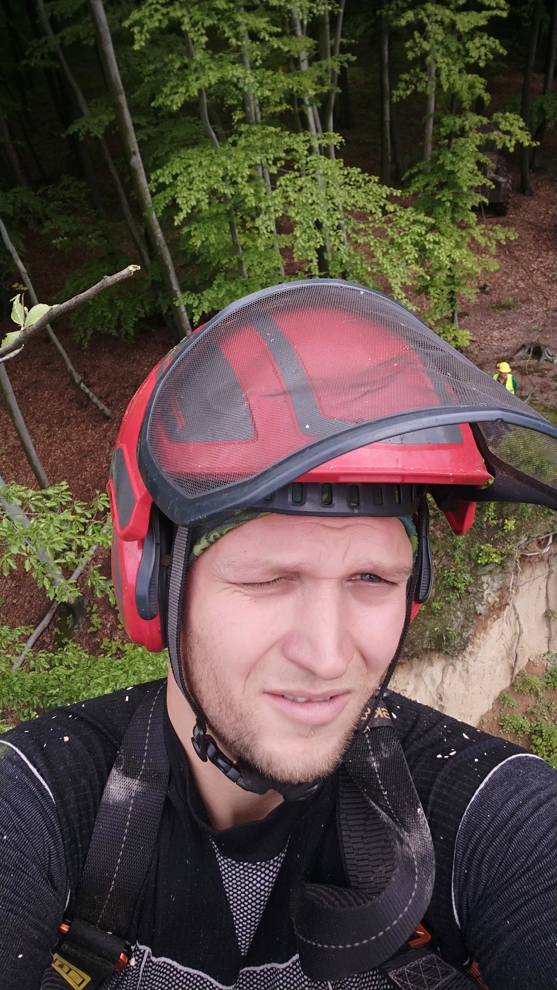 Mężczyzna w czerwonym kasku ochronnym z siatką, mruży jedno oko, w tle drzewa i drugi pracownik w oddali.