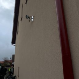 DEVELOPMENT SP Z O O - Jasna elewacja budynku z zamontowanym reflektorem i czerwoną rynną. Widok z dołu, na tle pochmurnego nieba i fragmentu zabudowy.