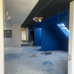 DEVELOPMENT SP Z O O - Wnętrze w trakcie malowania: ściany w odcieniach błękitu i szarości, czarny sufit. Malarz na drabinie, widoczne narzędzia i kable. Skos dachu z oknem.