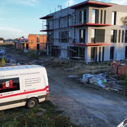 DEVELOPMENT SP Z O O - Wznoszony budynek wielorodzinny z szarymi elewacjami i czerwonymi akcentami, widoczny z perspektywy placu budowy z zaparkowanym busem firmy remontowej.