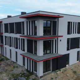 Nowoczesny, trzypiętrowy budynek z szarą elewacją i czarnymi, pionowymi panelami. Czerwone akcenty na daszkach balkonów. Widok z góry, teren budowy w tle.