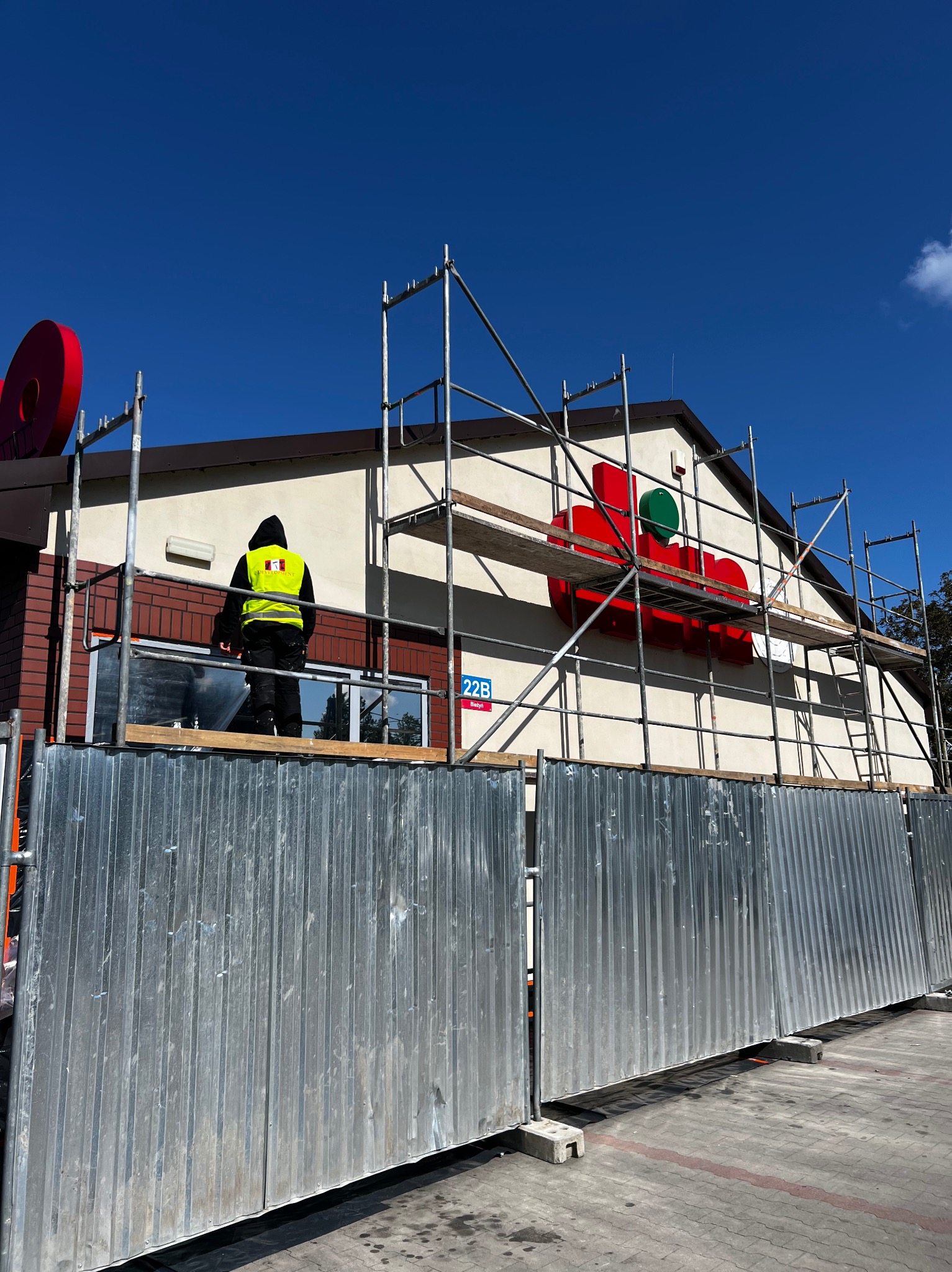 Pracownik w kamizelce na rusztowaniu, montuje logo na elewacji budynku. Widoczne metalowe ogrodzenie i fragment elewacji z cegły.
