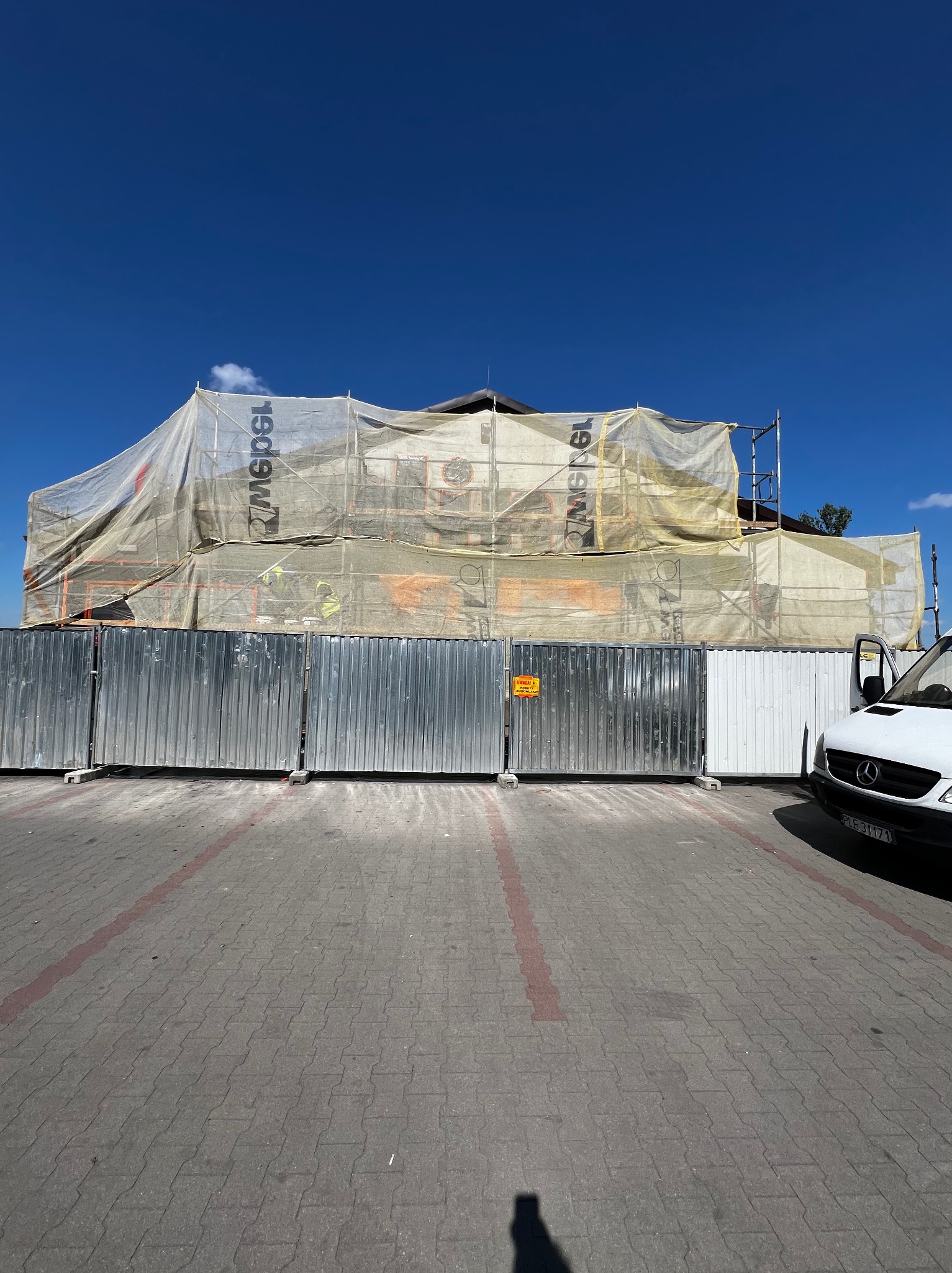 Budynek w trakcie prac elewacyjnych, osłonięty siatką i ogrodzony metalowym płotem, widoczny fragment białego samochodu i błękitne niebo.