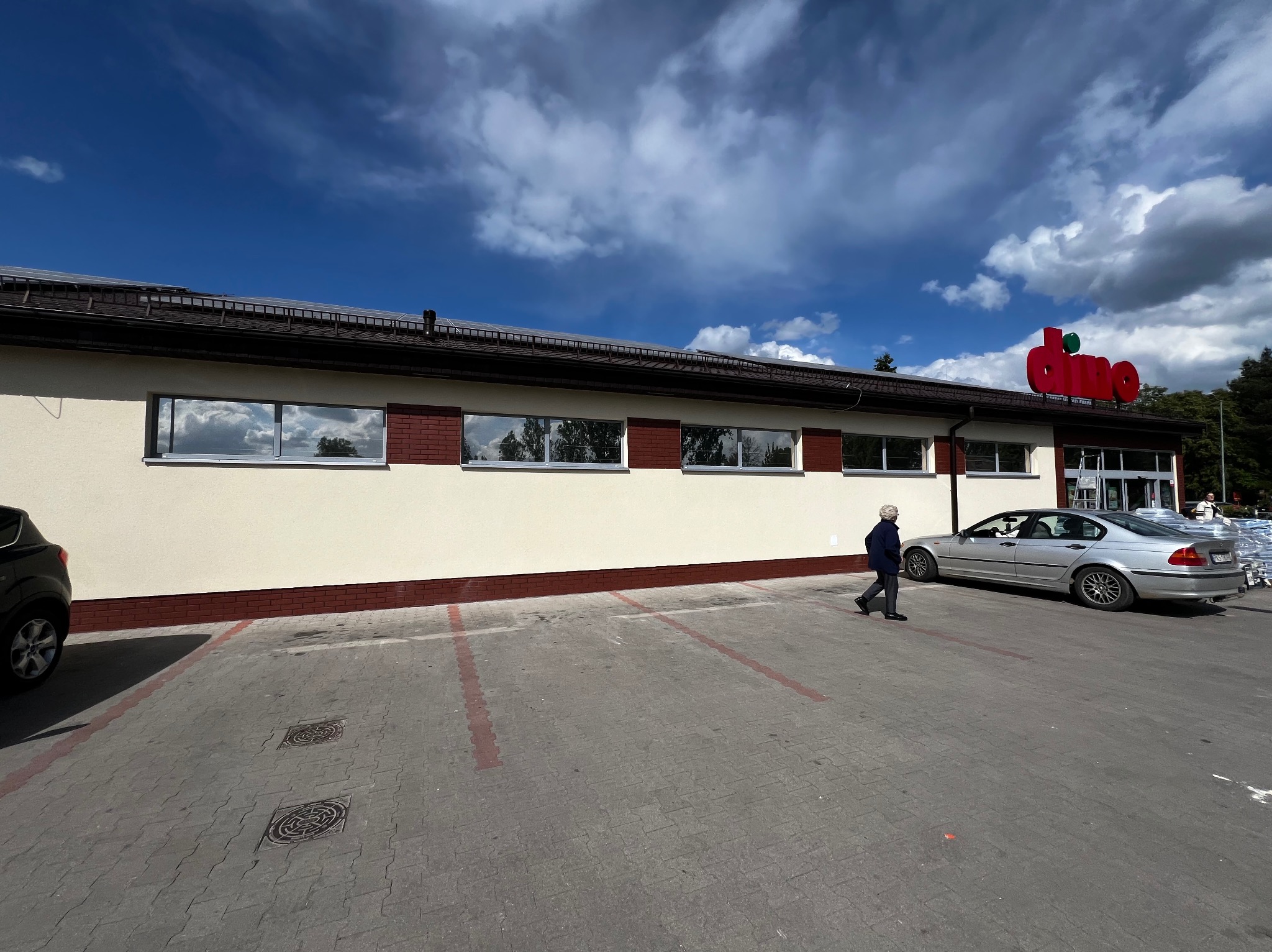 Budynek sklepu Dino z kremową elewacją, brązowymi detalami i logo na dachu. Na parkingu stoi srebrny samochód, a obok przechodzi starsza kobieta.