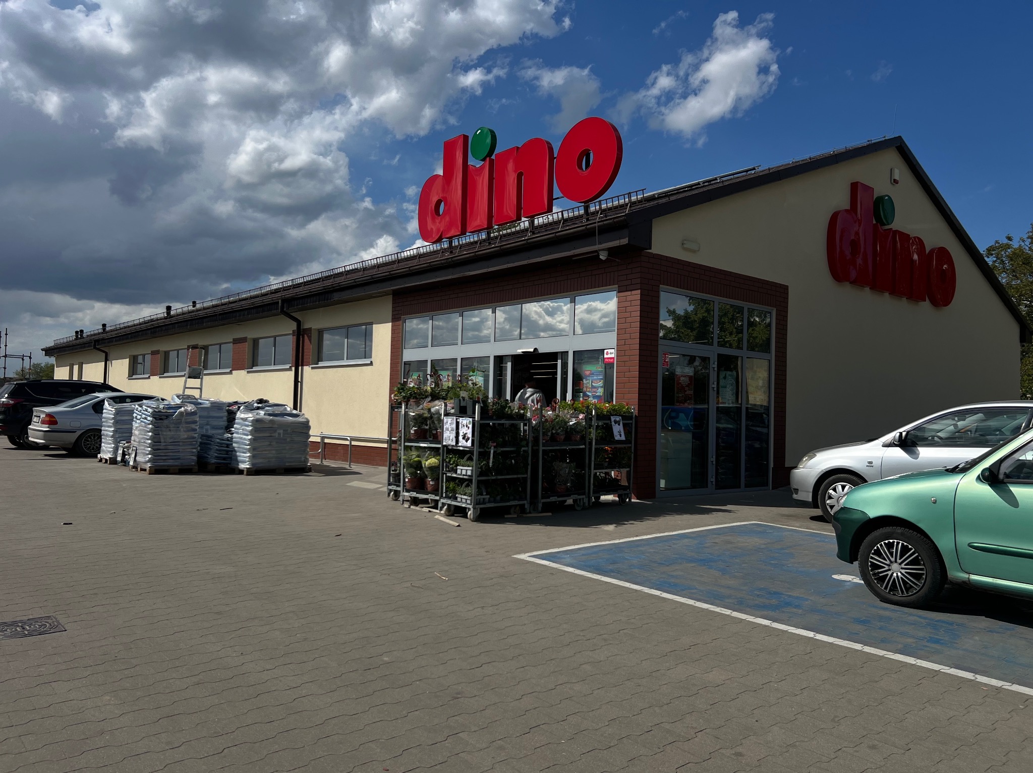 Fasada budynku sklepu Dino z czerwonym logo na tle błękitnego nieba z chmurami. Na parkingu zaparkowane samochody i palety z towarem.