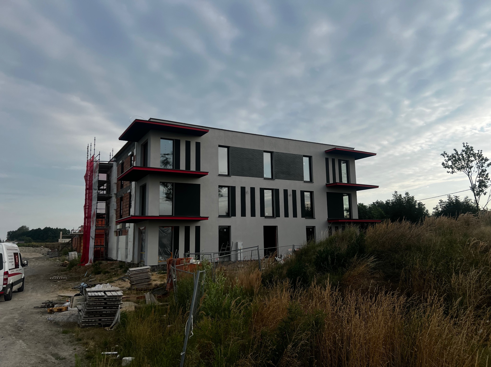 Nowoczesny, trzypiętrowy dom w trakcie budowy, z szarymi ścianami i czerwonymi akcentami, otoczony trawą i rusztowaniami. Widoczny samochód dostawczy.