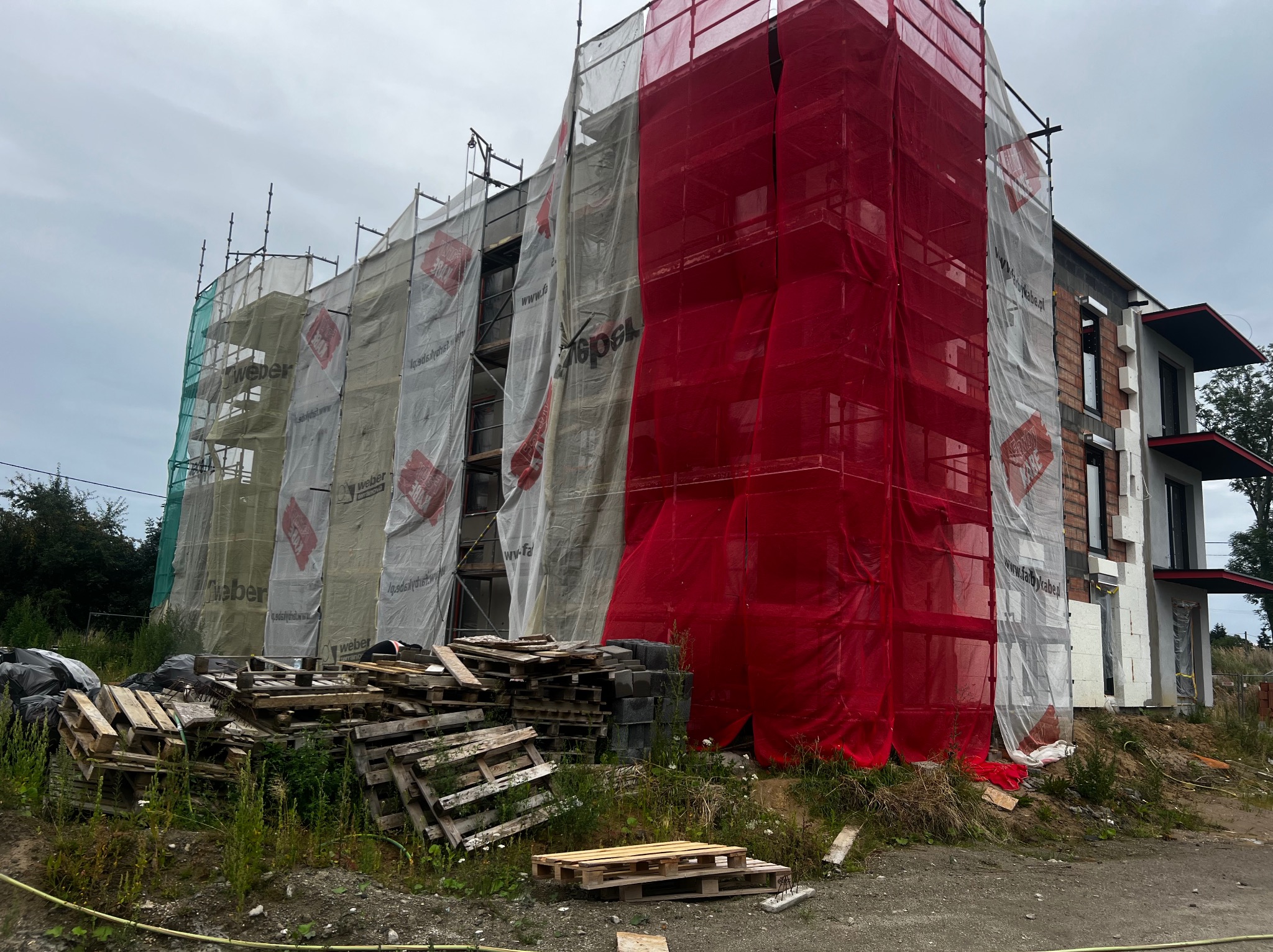 Budynek w trakcie ocieplania elewacji, osłonięty siatką ochronną z reklamami, widoczne rusztowania. Palety i materiały budowlane na pierwszym planie.