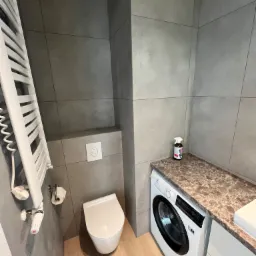 Nowoczesna łazienka z szarymi, kwadratowymi płytkami, podwieszana toaleta, biały grzejnik drabinkowy, pralka pod blatem imitującym marmur i butelka środka czyszczącego.