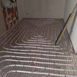 Białe rury ogrzewania podłogowego ułożone na folii z czerwoną siatką, widoczny poziomica oparta o ścianę, surowy stan wykończenia pomieszczenia.