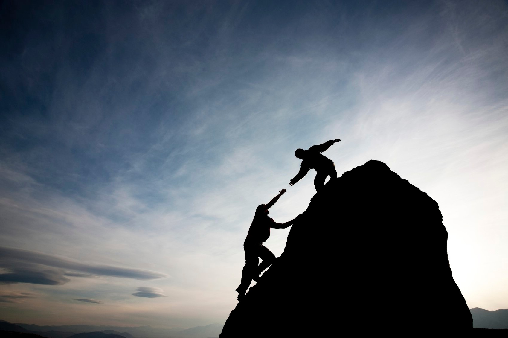 Silhouettes of two people climbing a rocky peak against a cloudy sky; one extends a hand to help the other, symbolizing support and overcoming challenges.
