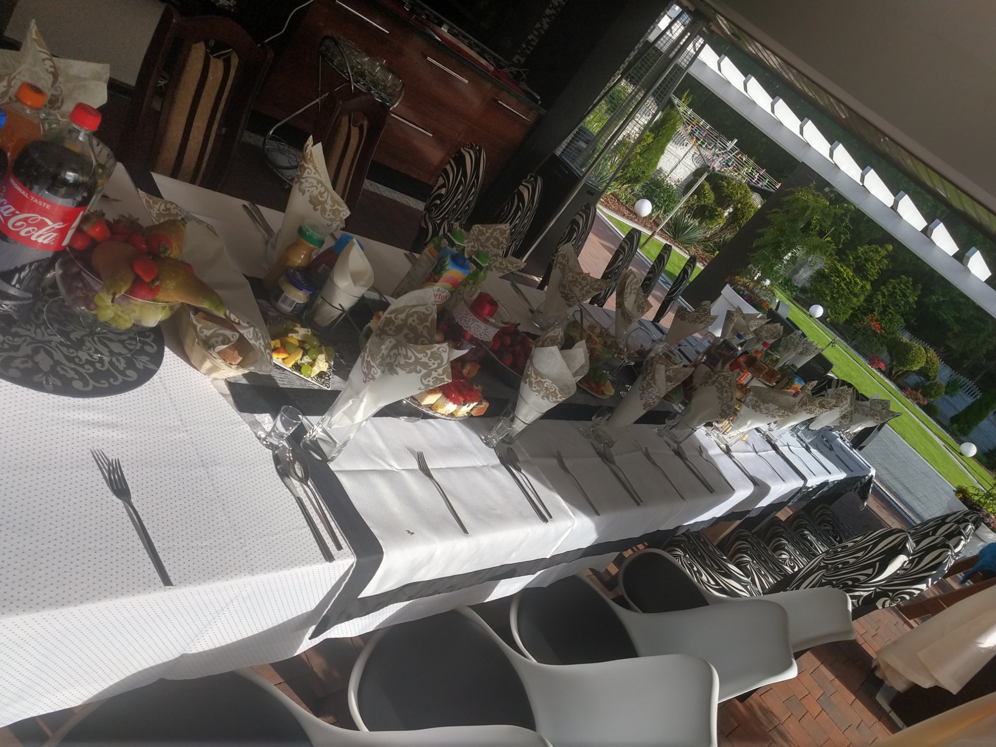 Długi stół z białym obrusem, nakryty do posiłku na zewnątrz, z dekoracjami, owocami, napojami i sztućcami, otoczony krzesłami w dwóch stylach: nowoczesne białe krzesła i krzesła z wzorem zebry,...