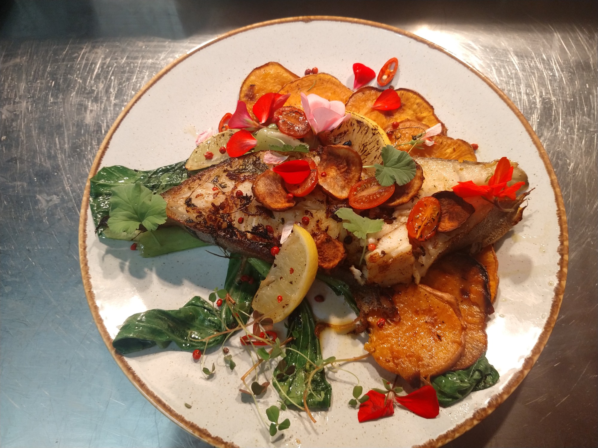 Elegancko podane danie z ryby z grillowanymi batatami, liśćmi szpinaku, plasterkami cytryny i jadalnymi kwiatami na talerzu z kamionki.